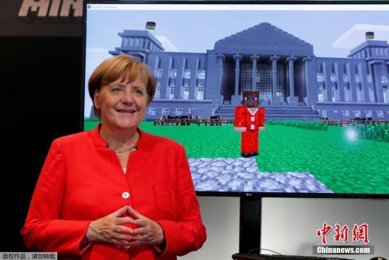 默克尔站在一台显示屏旁，屏幕中是游戏《MineCraft》的画面，其中还有以默克尔为原型制作的游戏角色，站在德国国会大楼前，如同现实中的场景。