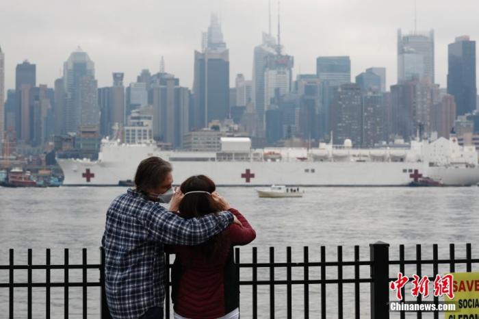 当地时间3月30日，美军医疗舰“安慰号”航行抵达纽约曼哈顿，新泽西威霍肯两位民众戴着口罩相拥眺望“安慰号”进入码头。中新社记者 廖攀 摄