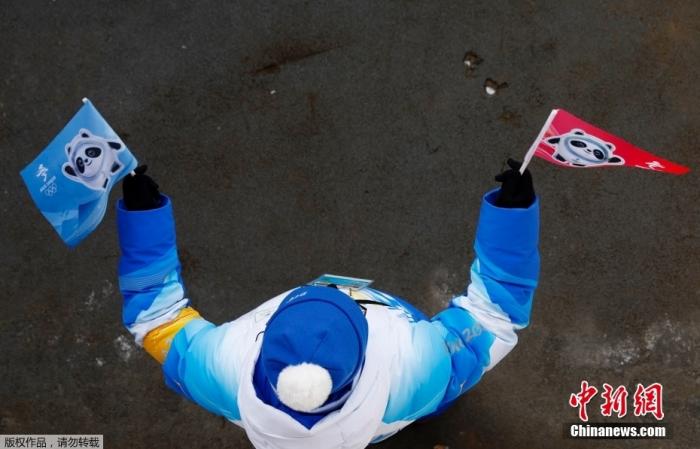 2月6日，冬奥会高山滑雪男子滑降赛场，志愿者手持“冰墩墩”旗子在场边为运动员加油。