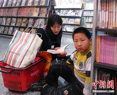 1月19日，北京某书店一位心不在焉的小朋友被记者吸引。寒假期间春节将至，有些心急的家长想利用假期为孩子补课。图中一位母亲正在为孩子读书中的内容，不想孩子被记者的相机快门声吸引。中新社记者 徐冬冬 摄